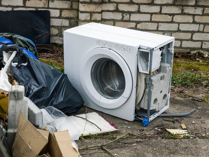 Image of white broken washing machine outdoors next to the garbage disposal.