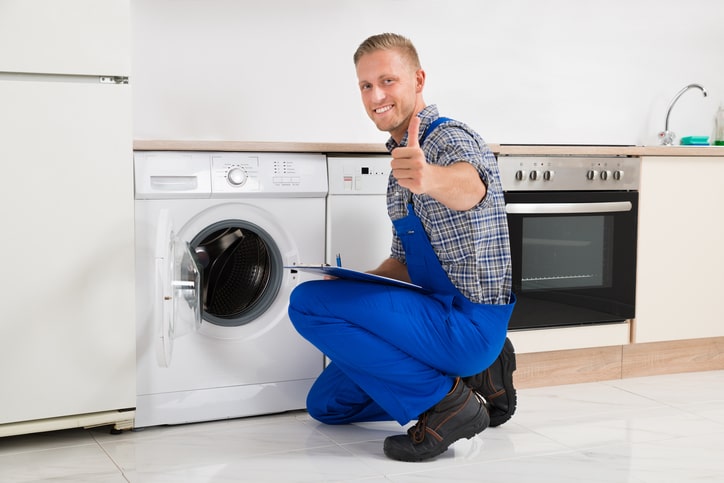 Man staring at the camera with his thumb up while in the background he has a washing machine, dishwasher, fridge and freezer and a stove.